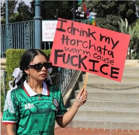 Indigenous woman holding a sign that says "I drink my horchata warm, because fuck ICE", while standing outside of a stairway located outdoors.  The picture is taken during daylight hours.